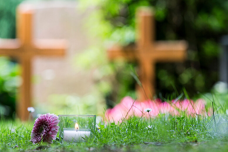 Zwei unscharfe Holzkreuze im Hintergrund. Davor steht eine brennende Kerze und eine lilafarbene Blume liegt links in der Wiese daneben.